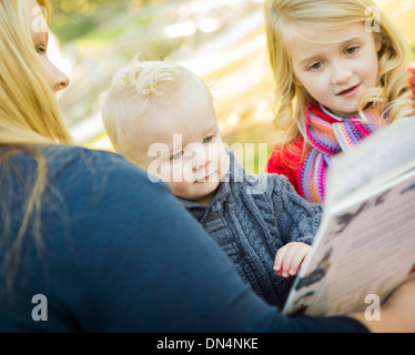 Mother Reading a Book to Her Two Adorable Blonde Children Wearing Winter Coats Outdoors. Stock Photo