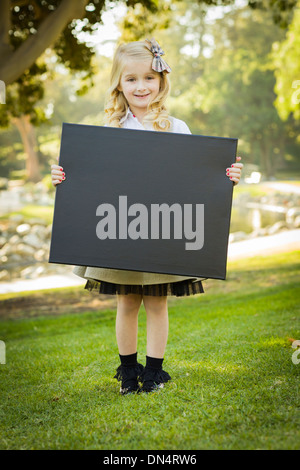 Cute Little Blonde Girl with a Bow in Her Hair Holding a Black Chalkboard Outdoors. Stock Photo