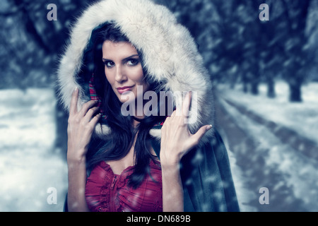 Woman in cape with hood standing on wooded path in the snow Stock Photo