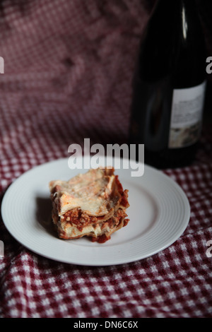 A slice of homemade meat lasagna on a white plate with a bottle of red wine in the background on a checkered table cloth Stock Photo