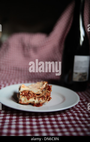 A slice of homemade meat lasagna on a white plate with a bottle of red wine in the background on a checkered table cloth Stock Photo