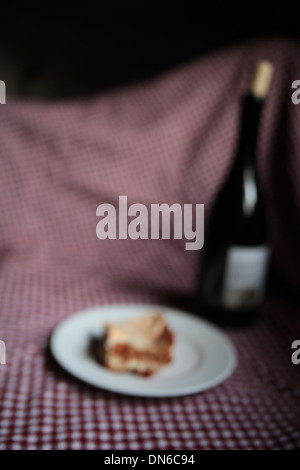 A slice of homemade meat lasagna on a white plate with a bottle of red wine in the background on a checkered table cloth Stock Photo