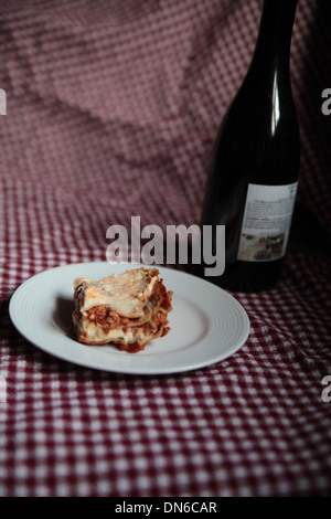 A slice of homemade meat lasagna on a white plate with a bottle of red wine in the background on a checkered table cloth Stock Photo