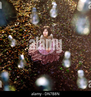 Woman with serious expression sitting on forest floor, surrounded by light bulbs / Drops Stock Photo