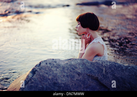 A thoughtful woman sits on a rock by a still pond in the forest, a ...