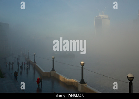 A woman in red coat caught in a patch of sunshine among figures walking in fog along Thames Stock Photo