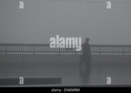 Silhouetted figure of a man in a hat and overcoat carrying a briefcase walking along the Thames in the fog Stock Photo