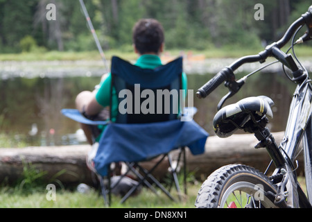 A teenage boy fishing by the lakeside in his camp chair. Focus on the bike. Stock Photo