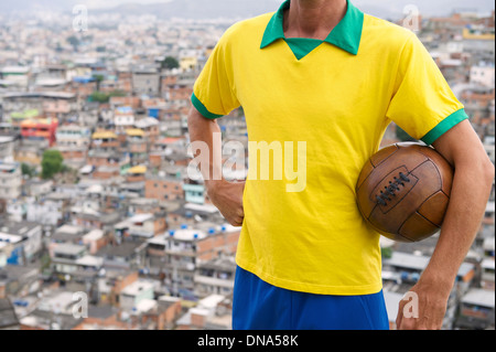 Brazilian football player standing in Brazil team colors holding vintage soccer ball favela background in Rio de Janeiro Brazil Stock Photo