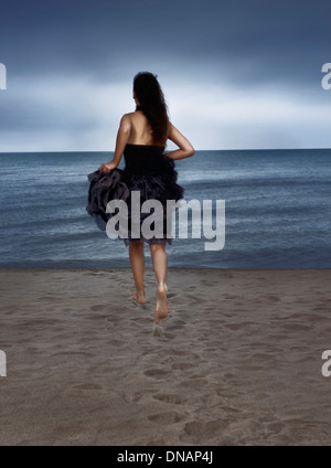 Young woman in black dress running towards the shore, rear view Stock Photo