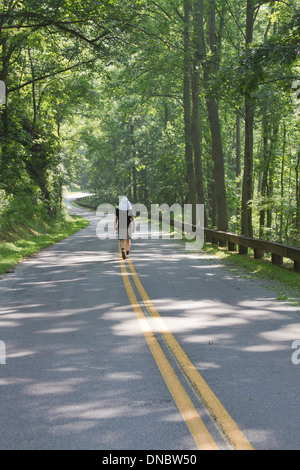 A man viewed from behind wearing a curtained hat walks down a long and winding road leading deep into the forest unknown Stock Photo