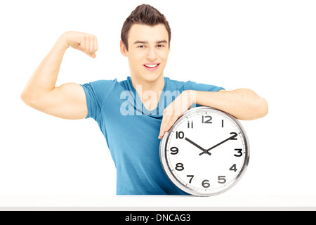 Young smiling man seated on a table with wall clock showing his muscles Stock Photo