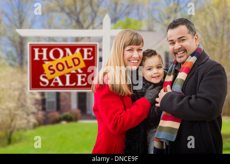 Warmly Dressed Young Mixed Race Family in Front of Sold Home For Sale Real Estate Sign and House.