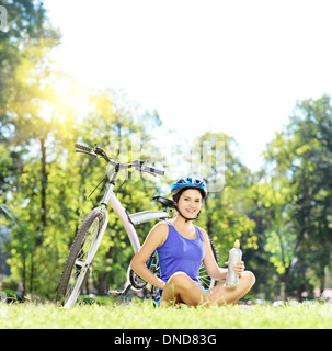 Young female biker sitting on a green grass next to a mountain bike and drinking water Stock Photo