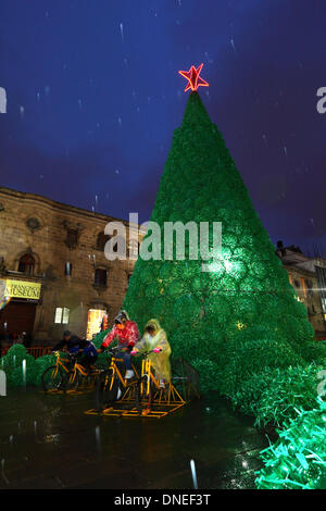 La Paz, Bolivia. 24th Dec, 2013. Locals pedal in the rain on bicycles connected to batteries to provide electrical power for the lights on an ecological Christmas tree in Plaza San Francisco. The tree is over 15m tall, contains approx. 50,000 recycled plastic drinks bottles and was a project conceived and planned by the La Paz City Government. Credit:  James Brunker / Alamy Live News Stock Photo