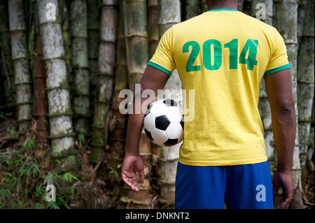 Brazilian soccer player in 2014 shirt standing in front of tropical bamboo jungle background holding football Stock Photo