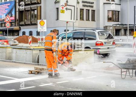 Two roadworkers in orange signal coloured workwear, one man with hammer, one man cutting a stone block with a angle grinder Stock Photo