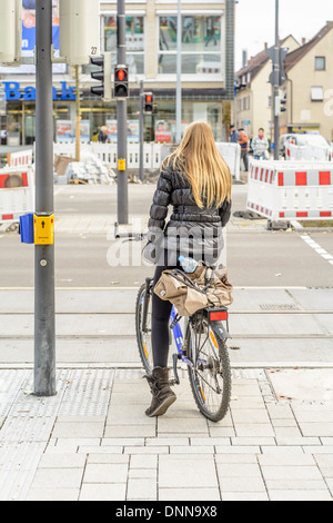 Young Caucasian woman with long blond hair, wearing a quilted jacket, riding a bicycle, waiting at red traffic lights Stock Photo
