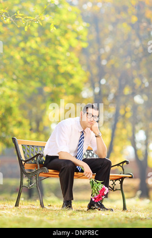 Sad young man holding a bouquet of flowers and sitting on a wooden bench in a park Stock Photo