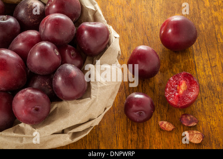 An image of red plums in a crumpled brown paper bag on a wood table. Stock Photo