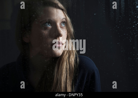 Portrait of young woman behind window, wet with raindrops, looking up Stock Photo