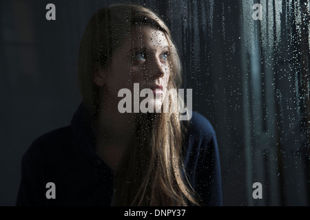 Portrait of young woman behind window, wet with raindrops, looking up Stock Photo