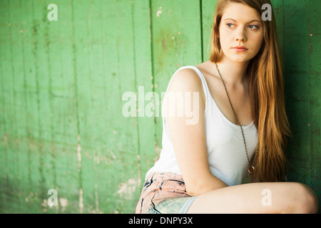 Portrait of young woman outdoors, crouching down by wall, looking to the side Stock Photo