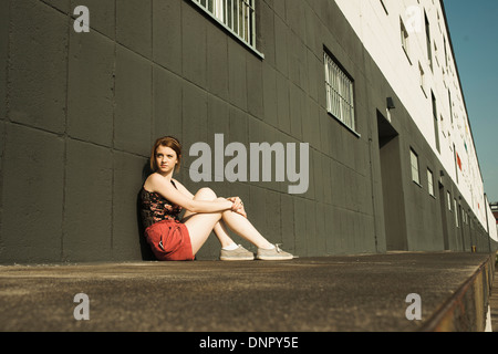 Teenage girl sitting and leaning against wall, looking away into the distance Stock Photo