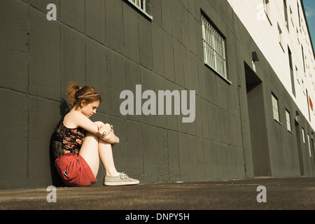 Teenage girl sitting on ground and leaning against wall, looking downwards Stock Photo