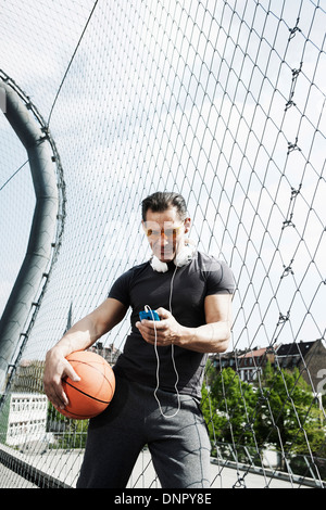 Mature man standing on outdoor basketball court holding basketball and looking at MP3 player, Germany Stock Photo