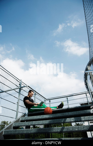 Mature man sitting at top of stairs on outdoor basketball court looking at tablet computer, Germany Stock Photo