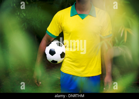 Brazilian football player in Brazil colors uniform standing in bamboo tropical jungle holding soccer ball Stock Photo