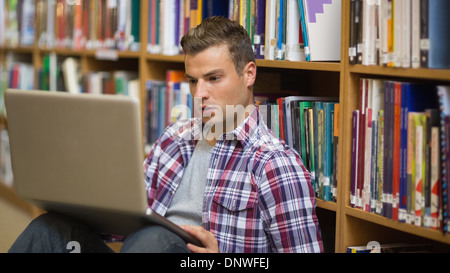 Serious young student sitting on library floor using laptop Stock Photo
