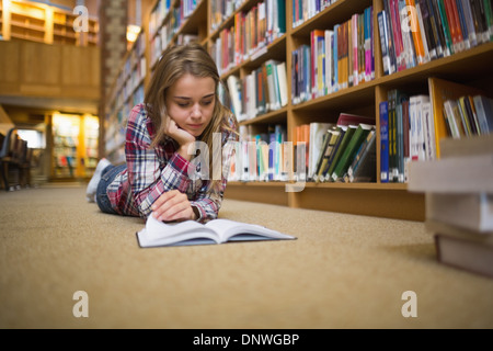 Pretty focused student lying on library floor reading book Stock Photo