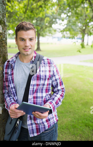 Handsome student leaning on tree looking at camera holding tablet Stock Photo