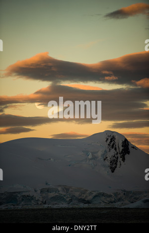 ANTARCTICA - The moon rises over a rocky mountain range in Paradise Harbor, Antarctica, while the setting sun casts a golden glow on the clouds and sky. Stock Photo