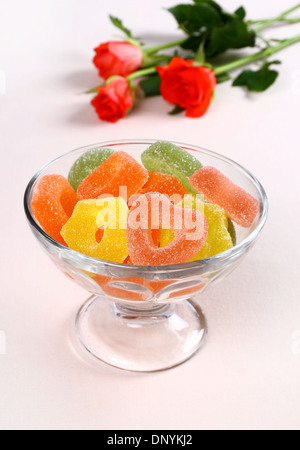 Colored candies with red hearts in glass bowl and roses in background Stock Photo