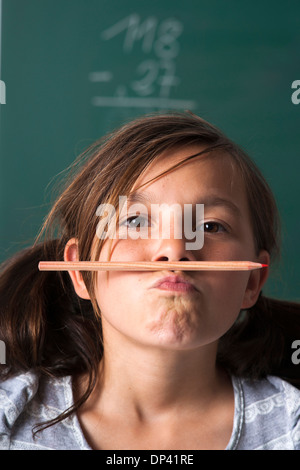 Portrait of girl standing in front of blackboard in classroom, holding pencil with mouth, Germany