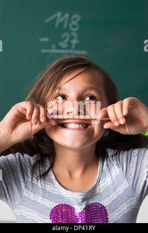 Portrait of girl standing in front of blackboard in classroom, holding pencil over mouth, Germany