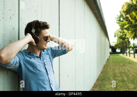 Young man standing outside leaning on wall of building, wearing headphones and sunglasses, Germany Stock Photo
