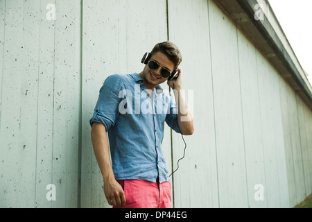 Young man standing next to wall of building outdoors, wearing headphones and sunglasses, Germany Stock Photo
