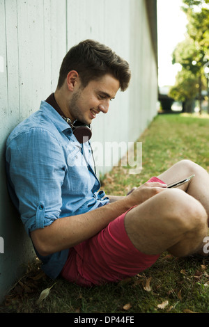 Young man sitting next to wall of building outdoors, with headphones around neck and looking at cell phone, Germany Stock Photo
