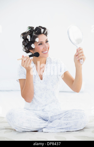 Cheerful brunette in hair rollers holding hand mirror and applying makeup Stock Photo