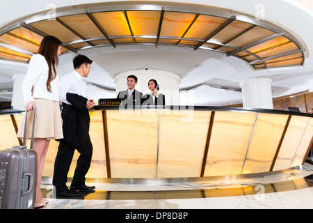 Asian Chinese woman and man arriving at front desk or reception of luxury hotel in business clothes with trolley Stock Photo