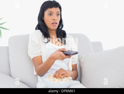 Shocked black haired woman in white clothes watching tv while eating popcorn Stock Photo