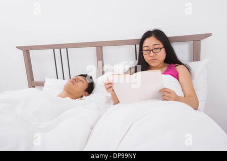 Asian young woman reading book by man sleeping in bed Stock Photo