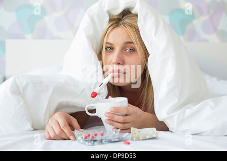 Portrait of sad woman with coffee mug taking temperature while wrapped in quilt on bed Stock Photo