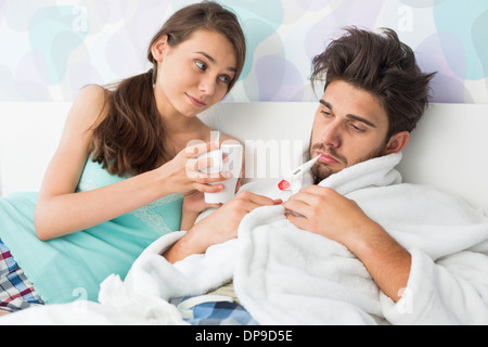 Young woman giving coffee mug to man with thermometer in mouth Stock Photo