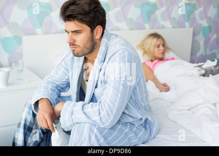 Sick man sitting on bed with woman sleeping in background at home