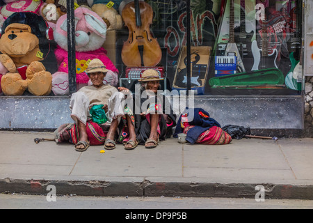 Indigenous man and woman begging while sitting on the sidewalk in front of a store window in Sucre, Bolivia. Stock Photo